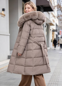A woman wearing a mid-length taupe puffer coat with a large brown faux-fur trimmed hood and a matching tie belt, standing on a city sidewalk.