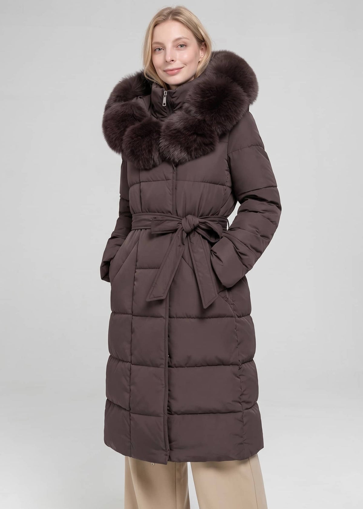 A woman wearing a long dark brown puffer coat with a large dark brown fur-trimmed hood and a matching waist tie, paired with beige pants, against a light gray studio background.