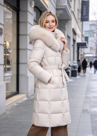A woman wearing a long cream-colored puffer coat with a large beige fur-trimmed hood and matching tie belt, standing on a city sidewalk.