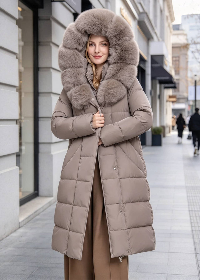 A woman wearing a long taupe-brown puffer coat with a large fluffy faux-fur trimmed hood, standing on a city sidewalk.