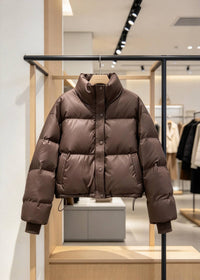 A brown puffer jacket with a high stand-up collar and button-front closure, hanging on a wooden hanger on a clothing rack inside a retail store.