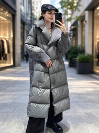 A woman takes a mirror selfie in an outdoor city setting, wearing a long, shiny silver-gray puffer coat, a black flat cap, black wide-leg trousers, and black boots.