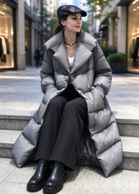 A woman sits on outdoor stone steps wearing a long silver-gray puffer coat, black wide-leg pants, black boots, and a black cap in an urban street setting.