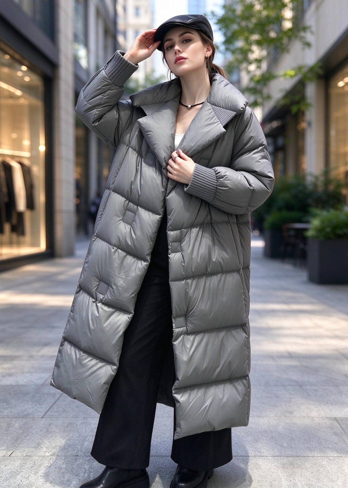 A woman wearing a long, oversized puffer coat in a medium gray color, styled with black pants and a black cap, standing on a city sidewalk.
