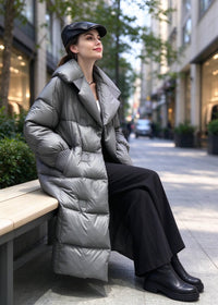 A woman sits on a bench in an urban street setting wearing a long, shiny silver-gray puffer coat, black wide-leg trousers, black ankle boots, and a black cap.