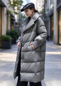 A woman wearing a long, oversized gray puffer coat with a high collar and black pants, standing on a city sidewalk. She is also wearing a black cap.