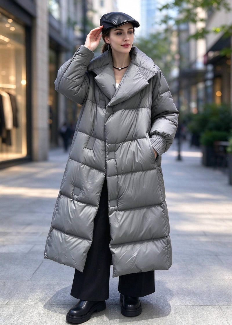 A woman stands on a city sidewalk wearing a long, shiny silver-gray puffer coat over a dark outfit, with a black cap and black shoes.