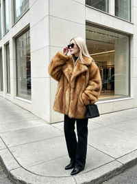 Blonde woman wearing a honey amber plush faux fox fur coat and sunglasses standing on a city street corner.