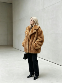 Blonde model in a honey amber plush faux fur jacket with black boots against a minimalist grey architectural wall.