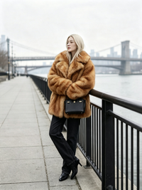 Woman leaning on a waterfront railing in a honey amber plush faux fur coat with a bridge in the background.