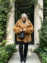 Blonde woman standing in front of an iron gate holding a black handbag, styled in a honey amber faux fur coat.