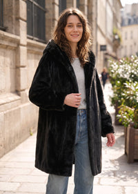 Front three-quarter outdoor view of a woman wearing a black hooded faux mink mid-length coat with the hood styled down.   