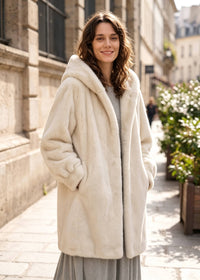 Full front three-quarter view of a woman in an off-white hooded faux mink mid-length coat with hands tucked in side pockets.   