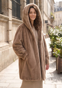 Side-front three-quarter street shot of a woman wearing a warm beige taupe faux mink mid-length coat with the plush hood up.   