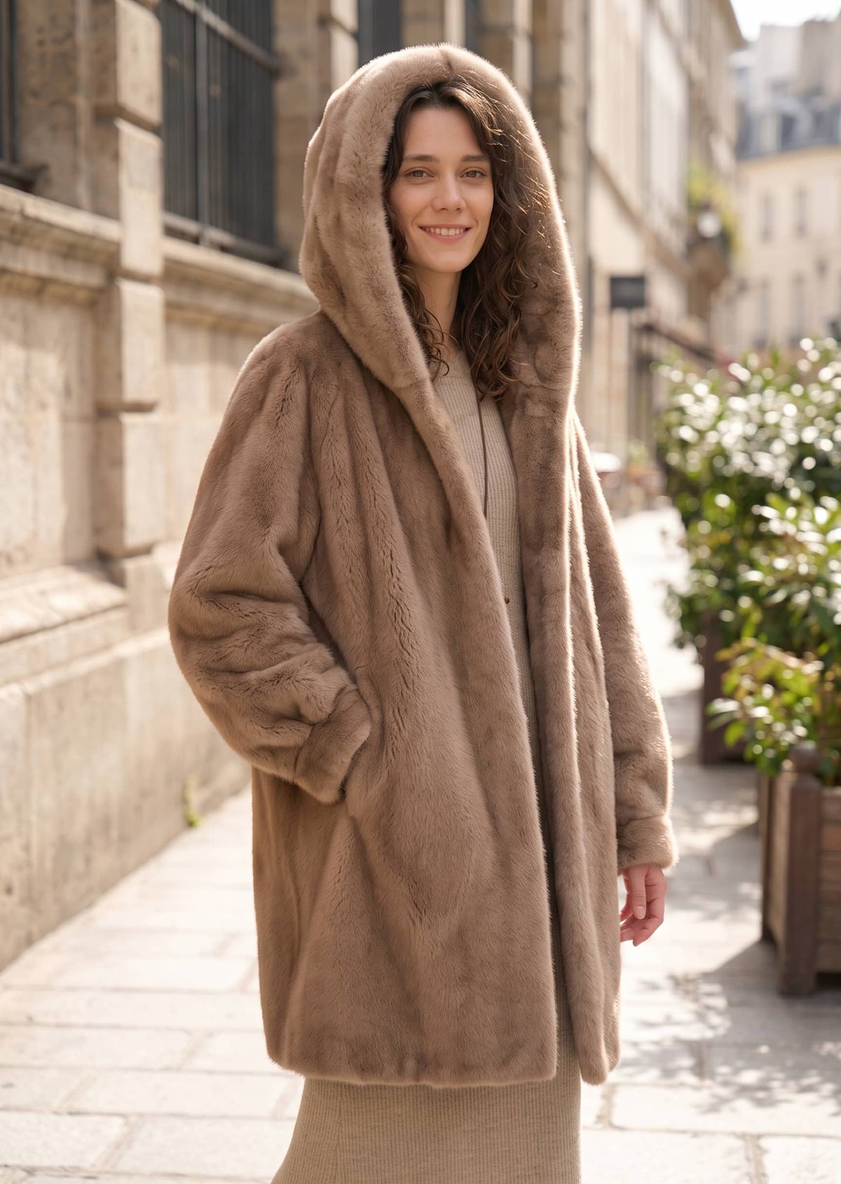 Side-front three-quarter street shot of a woman wearing a warm beige taupe faux mink mid-length coat with the plush hood up.   