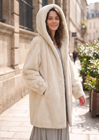 Three-quarter portrait of a woman wearing a winter off-white faux mink mid-length coat with the hood up on a city street.   
