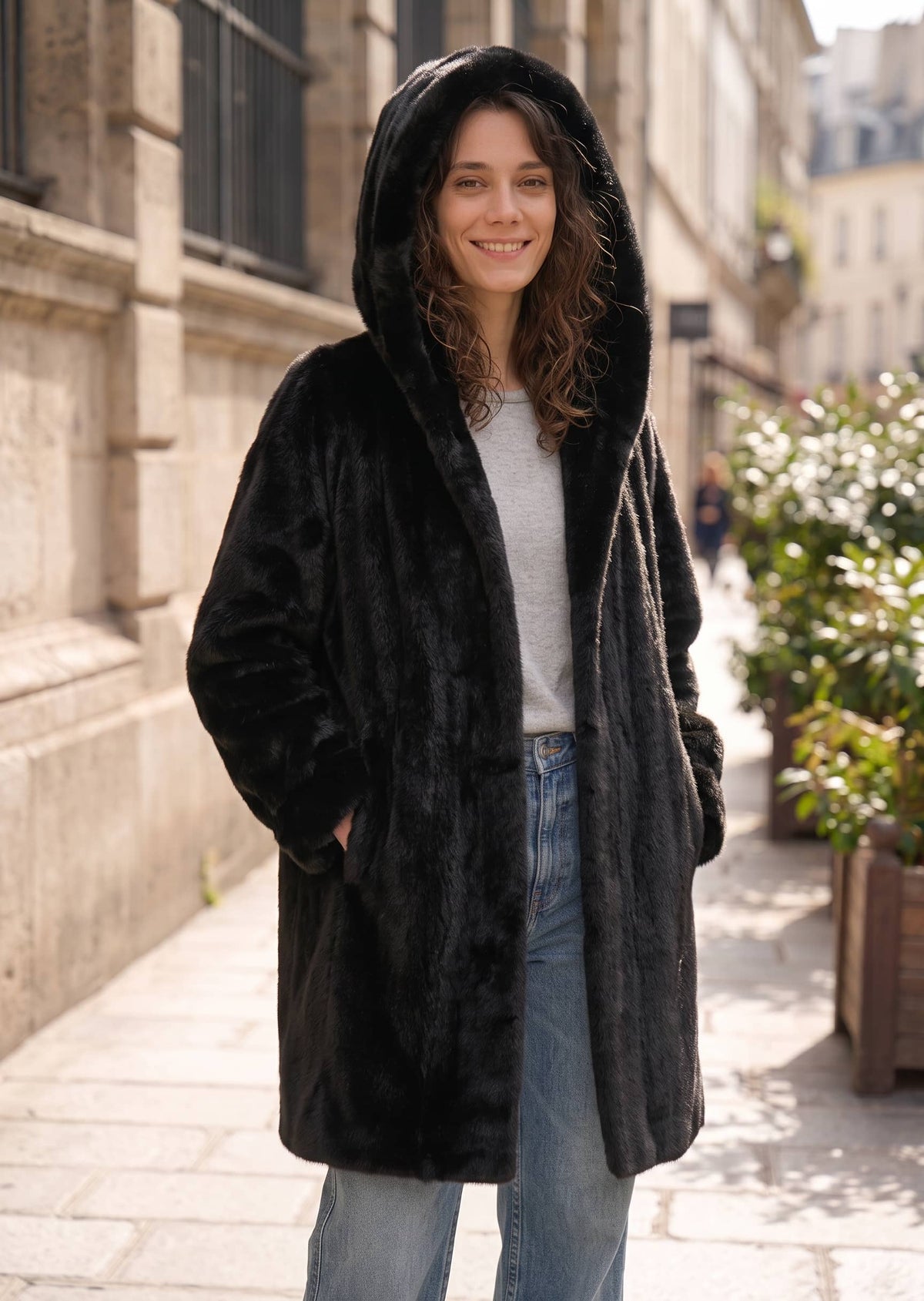 Front outdoor view of a woman wearing a black faux mink hooded coat with the hood up, styled with casual denim jeans.   