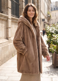 Side profile street photo of a woman wearing an oversized taupe brown hooded faux mink coat showing silhouette and sleeve volume.   
