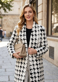 A woman standing on a city street wearing a long black and white houndstooth pattern coat over a black top and light gray pants, holding a beige handbag.