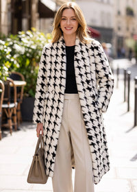 Front outdoor view of a smiling blonde woman wearing an open black and white houndstooth faux fur longline coat styled with wide-leg cream trousers.   