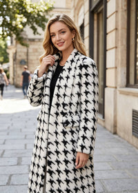 A woman wearing a long black and white houndstooth-patterned coat over a black top, standing on a stone sidewalk in front of a building.