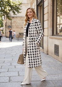 A woman walking on a city street wearing a long black and white houndstooth pattern coat over a black top and cream wide-leg trousers, carrying a beige handbag.