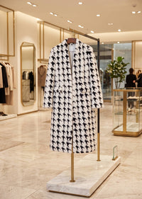 A long black and white houndstooth coat displayed on a gold-framed hanger stand inside a modern boutique with marble floors and warm lighting.