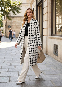 A woman walking on a city sidewalk wearing a long black-and-white houndstooth coat over a black top and cream wide-leg trousers, carrying a beige handbag.