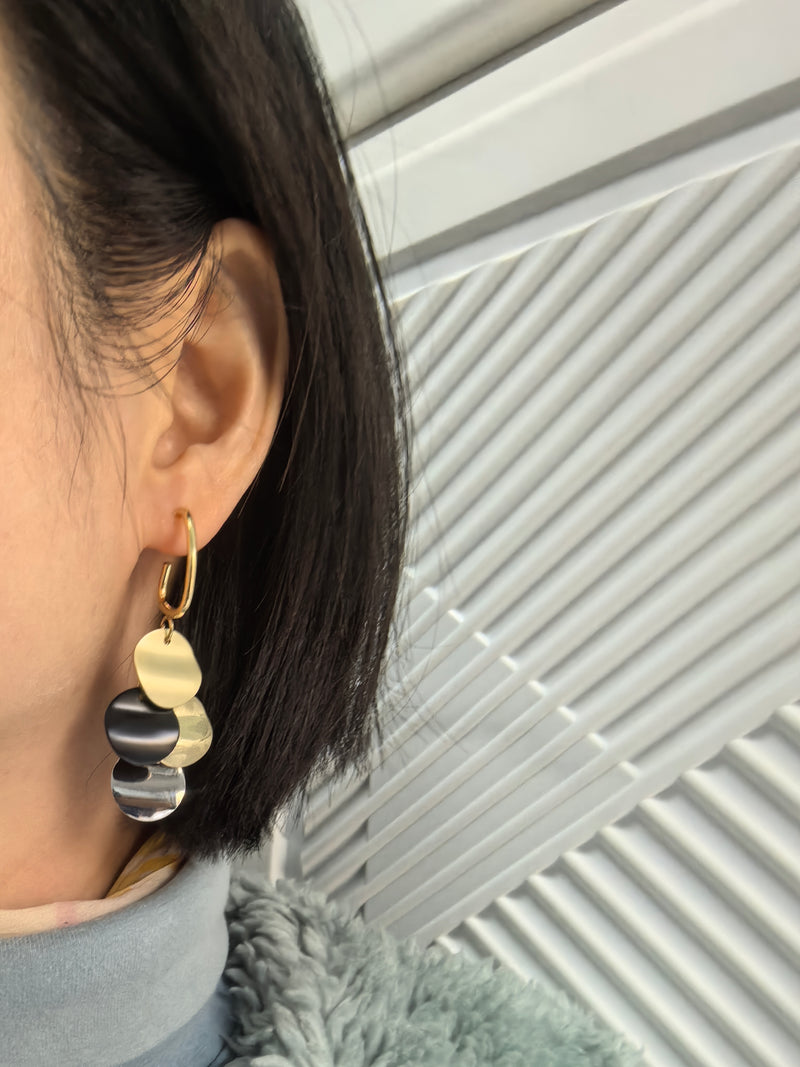 Close-up of an ear wearing a dangle earring with multiple small, round, multi-colored disks (gold, black, and silver/mother-of-pearl effect) hanging from a gold-tone hoop.