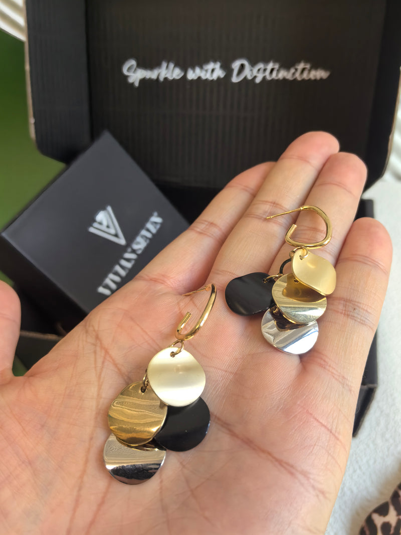 A hand holding a pair of multi-colored disc dangle earrings, with a black box labeled "Sparkle with Distinction" and "VIVIAN SEVEN" visible in the background.