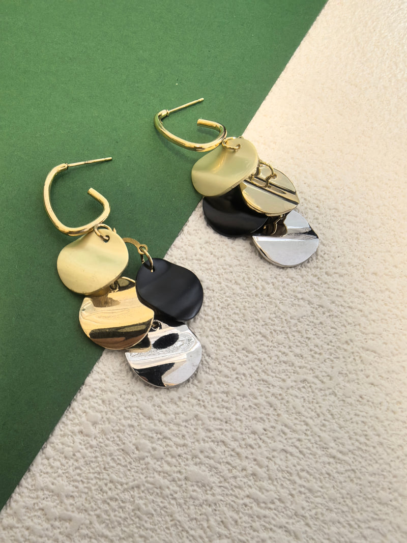 Pair of dangle earrings with round, multi-colored discs (gold, black, olive/light gold, silver/mother-of-pearl effect) displayed against a background of green and textured beige.