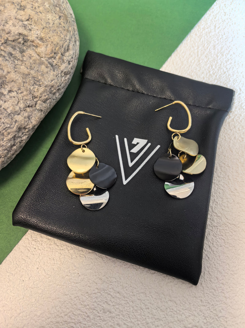 Pair of multi-colored disc dangle earrings resting on a small black pouch with a white "V" logo, next to a grey stone.
