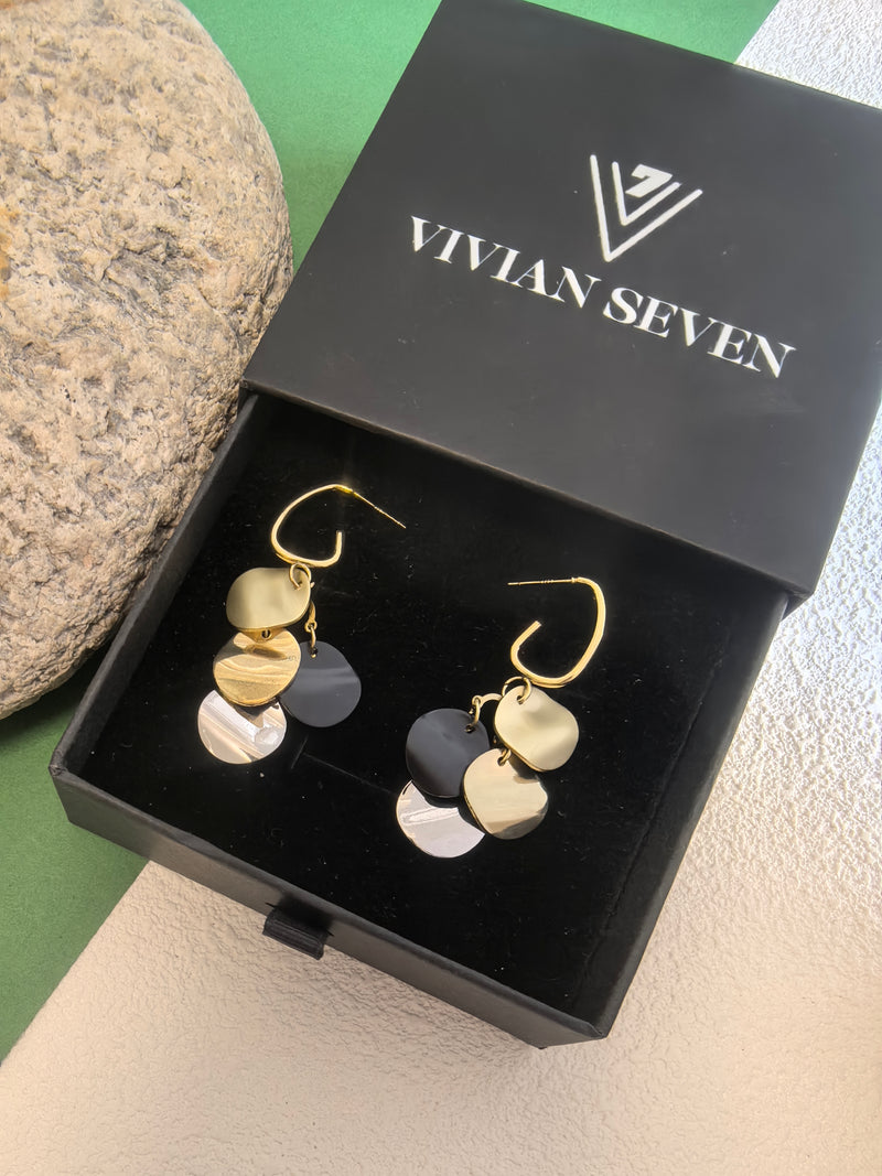Pair of multi-colored disc dangle earrings presented in an open black gift box labeled "VIVIAN SEVEN" on a textured surface next to a grey stone.