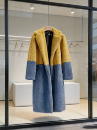 Women's mustard yellow and slate blue two-tone long faux fur coat hanging on a wooden hanger inside a store window display.