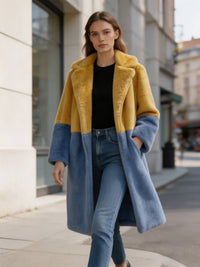 Model walking down a city street in a mustard yellow and slate blue two-tone long faux fur coat with blue jeans.