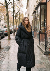 Vivian Seven black wrap down maxi coat with wide shawl collar and hood down, walking pose