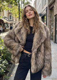 A woman stands on a city sidewalk wearing a voluminous shaggy faux-fur jacket in mixed brown, beige, and gray tones over a black top and black jeans.
