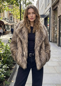 Front three-quarter street shot of a model with hands in pockets wearing a shaggy tipped brown faux fur wide-collar coat. 