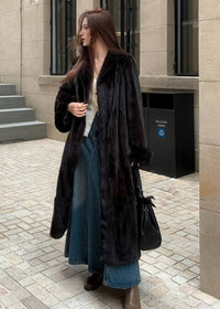 A side profile shot of the woman walking while wearing the black longline faux fur coat. The image shows the movement of the coat and the pairing with brown leather boots.