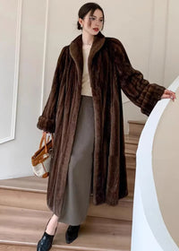 A full-length shot of a woman standing on wooden stairs wearing the brown longline faux fur coat. She is holding a tan and cream handbag and looking toward the camera.