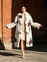 Front view of a model walking in a dramatic long cow print faux fur coat in front of a brick wall.