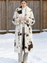 Woman standing in snow holding the lapels of a white and brown spotted faux fur winter long coat.
