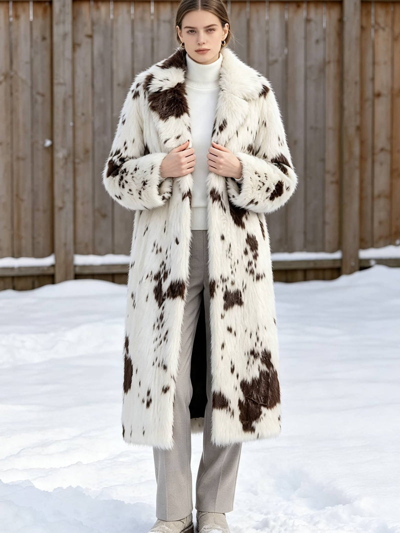 Woman standing in snow holding the lapels of a white and brown spotted faux fur winter long coat.