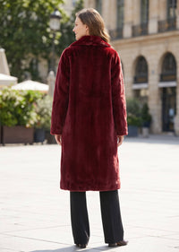 A woman viewed from behind wearing a long, plush burgundy faux fur coat over black wide-leg trousers, standing in an outdoor urban setting with a blurred stone building in the background.