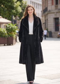 A woman stands on a city sidewalk wearing a long black faux-fur coat over a white top and black wide-leg trousers.