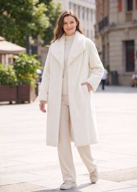 Full-body outdoor fashion shot of a woman modeling an off-white faux mink longline coat with matching monochrome pants.
