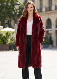 A woman wearing a long, plush burgundy faux fur coat over a white top and black pants, standing in an outdoor urban setting with blurred buildings and greenery in the background.