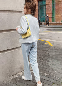 A full-length rear view of a young woman walking down a city sidewalk. She is wearing a light blue-gray textured tweed jacket, light-wash wide-leg jeans, white sneakers, and a small yellow shoulder bag.