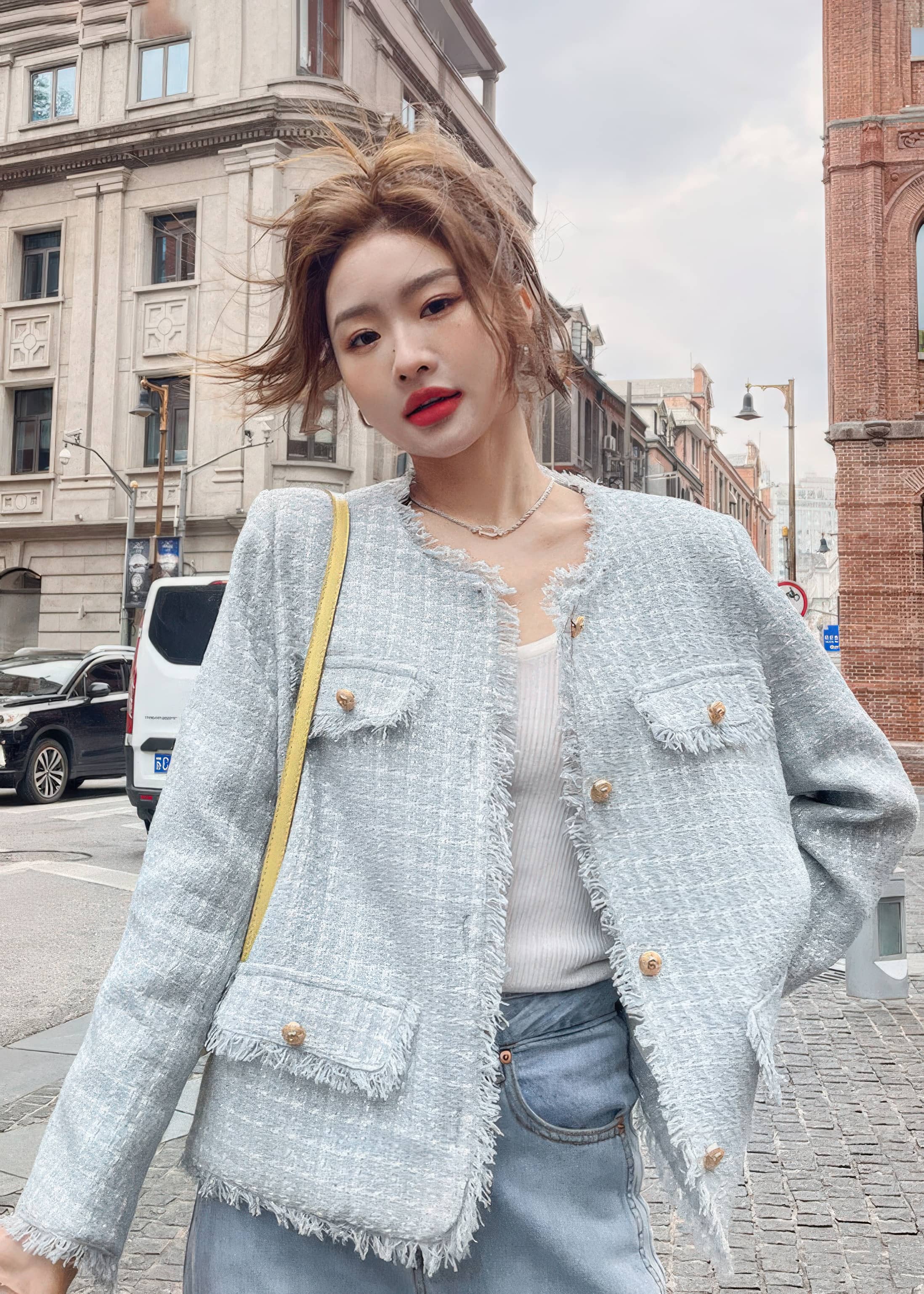 A medium close-up of a young woman with a messy updo posing on a city street. She wears an open, light blue-gray tweed jacket with raw-edge detailing and gold buttons over a ribbed white top.