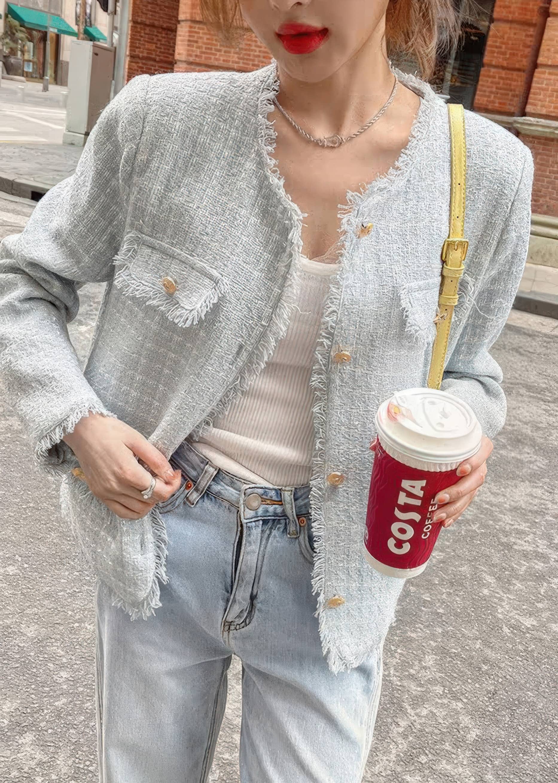 A close-up shot focusing on the details of a light blue-gray tweed jacket with prominent frayed edges and gold buttons. The person wearing it holds a red coffee cup and wears light-wash jeans.